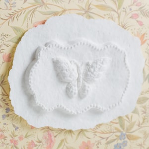 Op de afbeelding: Witte gipsen plaquette met een reli&euml;f van een vlinder. De vlinder bevindt zich in het midden van de plaquette en heeft een gedetailleerd ontwerp.