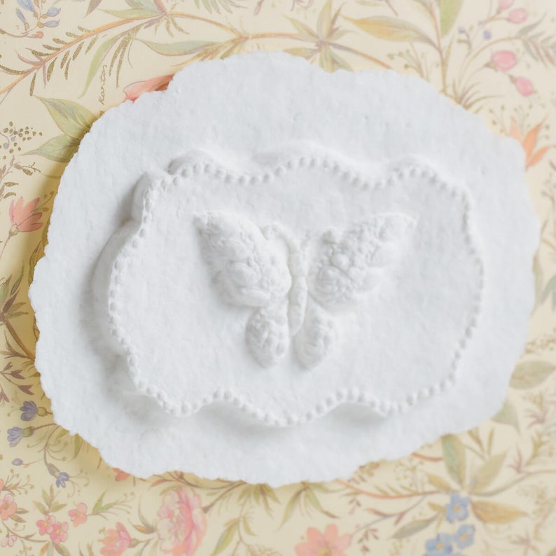 Op de afbeelding: Witte gipsen plaquette met een reli&euml;f van een vlinder. De vlinder bevindt zich in het midden van de plaquette en heeft een gedetailleerd ontwerp.