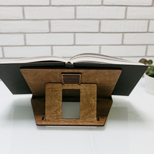 May include: A wooden book stand with a black book resting on top. The stand has a rectangular shape with a small opening in the back for ventilation.