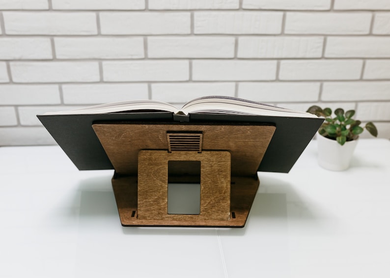 May include: A wooden book stand with a black book resting on top. The stand has a rectangular shape with a small opening in the back for ventilation.