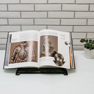 May include: An open book with illustrations of armor and helmets on a white surface. The book is on a black stand.