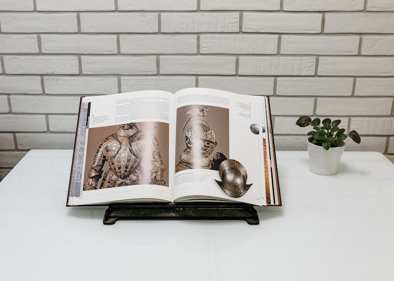 May include: An open book with illustrations of armor and helmets on a white surface. The book is on a black stand.