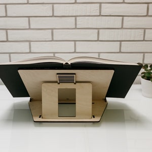 May include: A wooden book stand with a black book resting on top. The stand has a rectangular opening in the front and a small slatted opening on the top.
