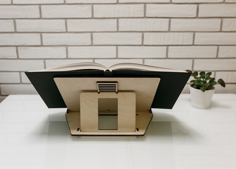 May include: A wooden book stand with a black book resting on top. The stand has a rectangular opening in the front and a small slatted opening on the top.