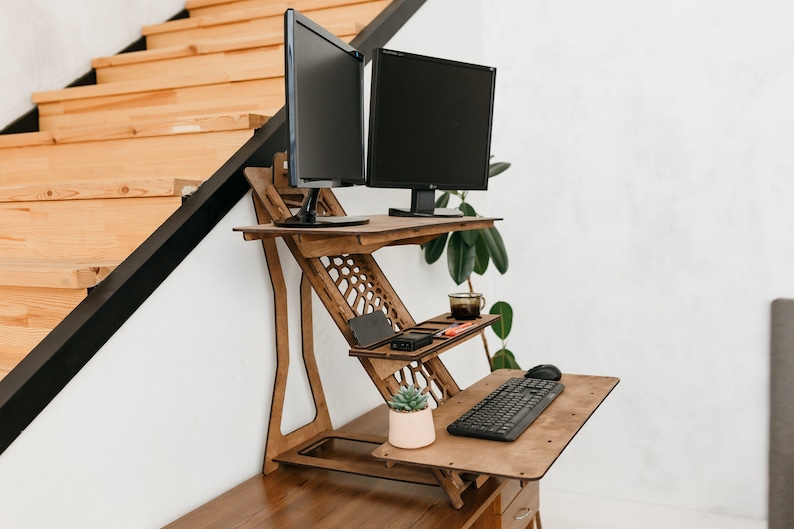 K&ouml;nnte beinhalten: Ein Stehpult aus Holz mit zwei schwarzen Computermonitoren, einer Tastatur, einer Maus, einer Topfpflanze und einer Tasse auf einem Regal. Der Schreibtisch ist in einem Heimb&uuml;ro eingerichtet.