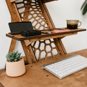 K&ouml;nnte beinhalten: Ein Stehpult aus Holz mit einem Wabenmuster. Der Schreibtisch hat zwei Ablagen, eine mit einem Telefon und einer kleinen schwarzen Box, die andere mit einer Tasse Kaffee und ein paar Gegenst&auml;nden. Eine Topfpflanze mit einer Sukkulente steht auf der unteren Ablage, und eine wei&szlig;e Tastatur und Maus befinden sich auf der Schreibtischoberfl&auml;che.