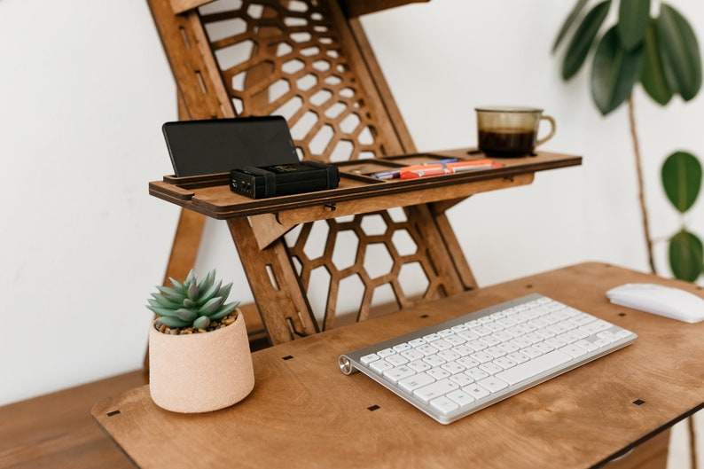 K&ouml;nnte beinhalten: Ein Stehpult aus Holz mit einem Wabenmuster. Der Schreibtisch hat zwei Ablagen, eine mit einem Telefon und einer kleinen schwarzen Box, die andere mit einer Tasse Kaffee und ein paar Gegenst&auml;nden. Eine Topfpflanze mit einer Sukkulente steht auf der unteren Ablage, und eine wei&szlig;e Tastatur und Maus befinden sich auf der Schreibtischoberfl&auml;che.