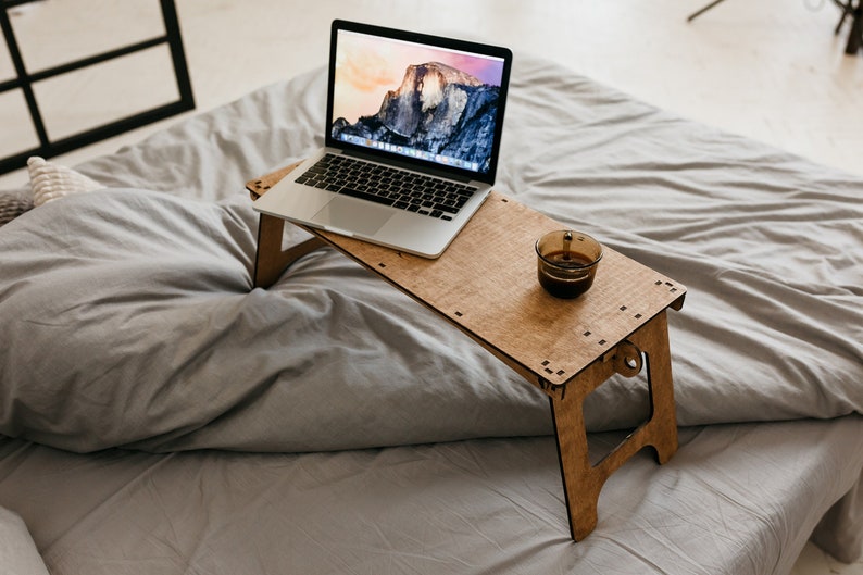May include: A wooden laptop stand with a laptop and a cup of coffee on it. The stand is on a gray bed.