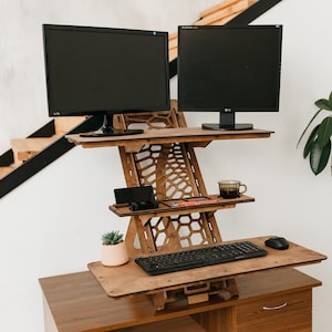 K&ouml;nnte beinhalten: Ein Stehpult aus Holz mit zwei schwarzen Computermonitoren, einer Tastatur, einer Maus, einer Tasse, einer Pflanze und einem Telefon auf der oberen Ablage. Das Pult ist auf einem Holzrahmen mit einem Wabenmuster erh&ouml;ht.