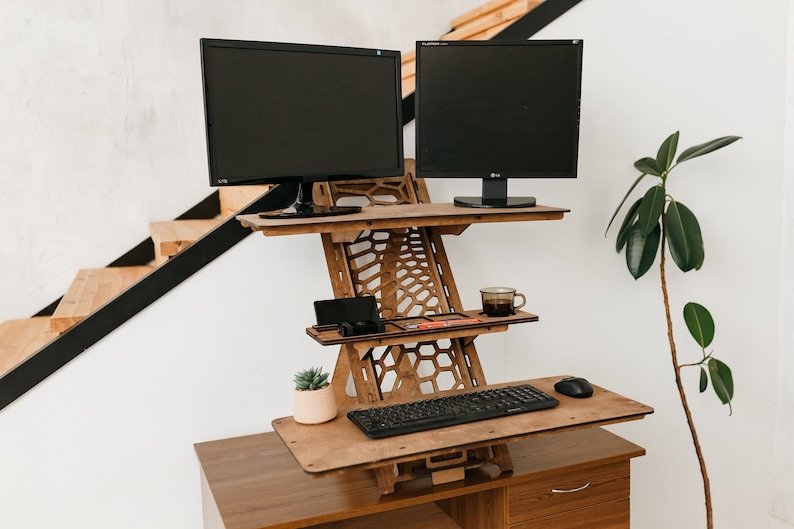 K&ouml;nnte beinhalten: Ein Stehpult aus Holz mit zwei schwarzen Computermonitoren, einer Tastatur, einer Maus, einer Tasse, einer Pflanze und einem Telefon auf der oberen Ablage. Das Pult ist auf einem Holzrahmen mit einem Wabenmuster erh&ouml;ht.
