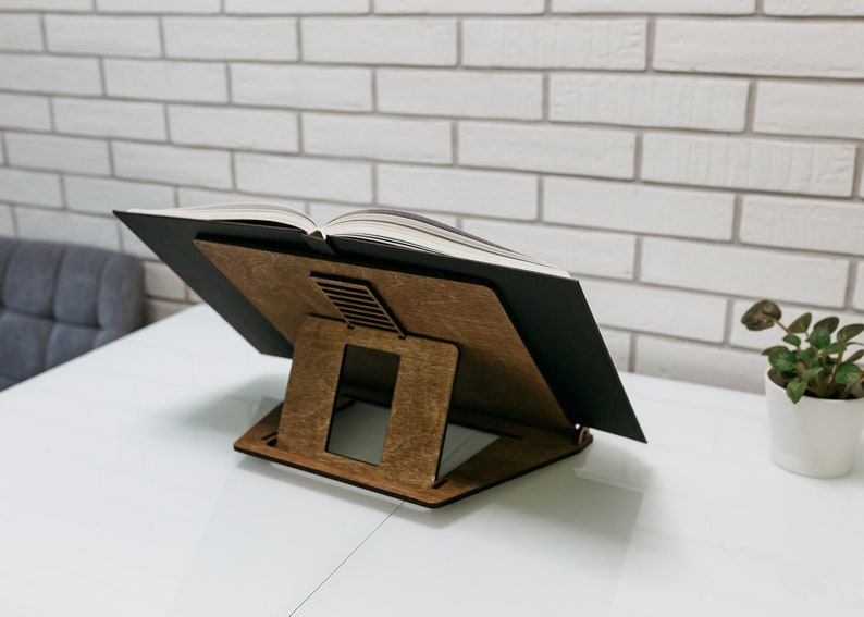 May include: A wooden book stand with a black book resting on top. The stand has a rectangular base and two angled supports. The book is open and the pages are visible.