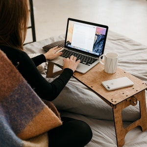 May include: A person working on a laptop computer on a wooden laptop stand. The laptop is open and the person is typing on the keyboard. A white mug and a white smartphone are on the stand. The person is sitting on a bed with a blanket.