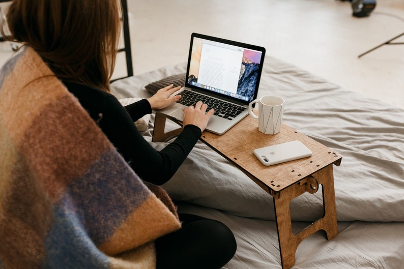 May include: A person working on a laptop computer on a wooden laptop stand. The laptop is open and the person is typing on the keyboard. A white mug and a white smartphone are on the stand. The person is sitting on a bed with a blanket.