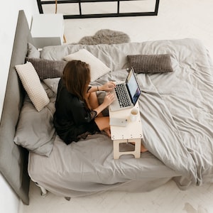 May include: A woman sits on a bed with a laptop on a wooden stand. She is wearing a black robe and has long brown hair. The bed has a gray duvet and white pillows. There is a rug with a geometric pattern on the floor.