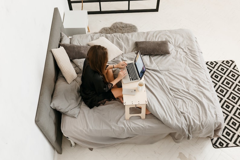 May include: A woman sits on a bed with a laptop on a wooden stand. She is wearing a black robe and has long brown hair. The bed has a gray duvet and white pillows. There is a rug with a geometric pattern on the floor.
