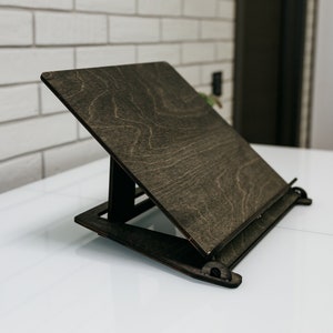 May include: A dark brown wooden book stand with a slanted surface and a metal bar to hold pages in place. The stand is made of plywood and has a natural wood grain.