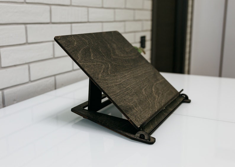 May include: A dark brown wooden book stand with a slanted surface and a metal bar to hold pages in place. The stand is made of plywood and has a natural wood grain.