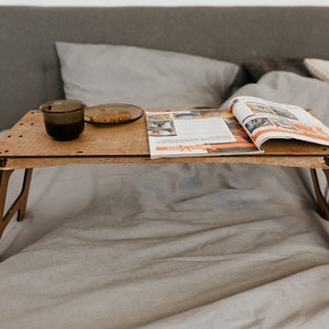 May include: A wooden folding tray table with a magazine and a cup and saucer on top. The tray table is sitting on a gray bed.