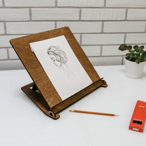 May include: A wooden easel with a drawing of a woman's face on a white sheet of paper. A pencil and a box of pencils are on a white surface.