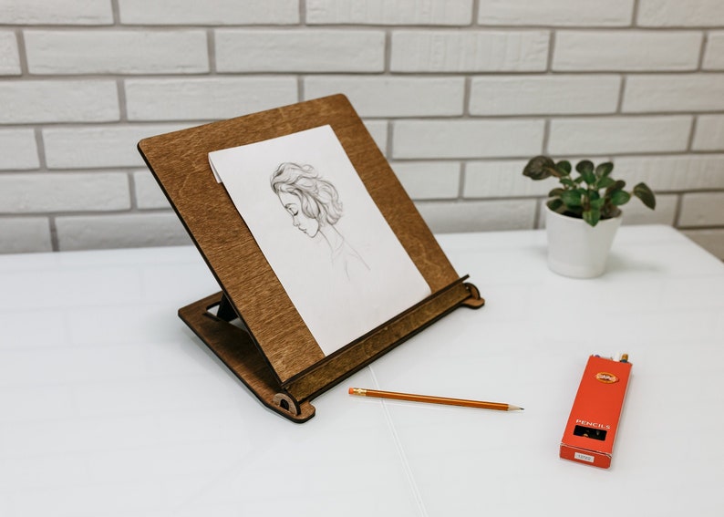 May include: A wooden easel with a drawing of a woman's face on a white sheet of paper. A pencil and a box of pencils are on a white surface.