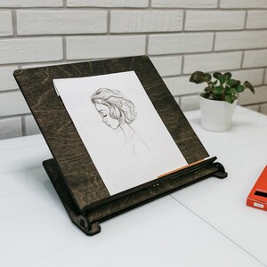 May include: A dark brown wooden easel with a drawing of a woman's face on a white sheet of paper. A box of pencils is on the white table surface.