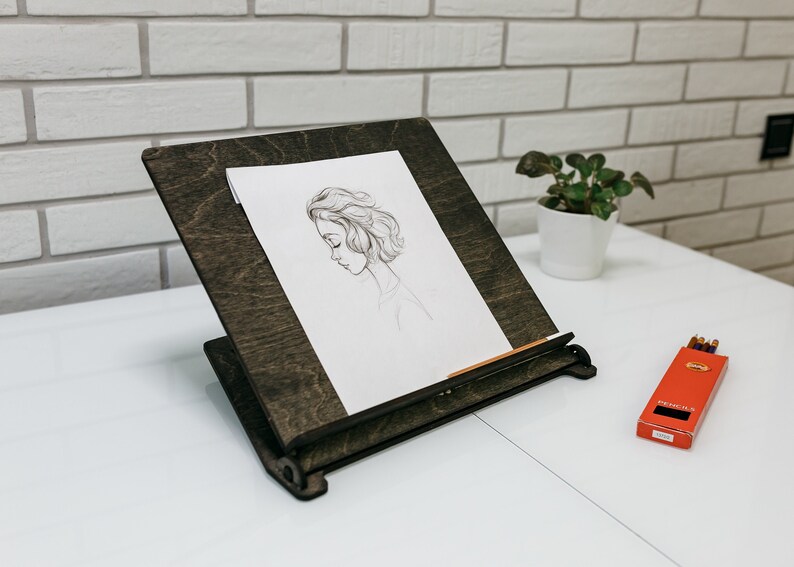 May include: A dark brown wooden easel with a drawing of a woman's face on a white sheet of paper. A box of pencils is on the white table surface.
