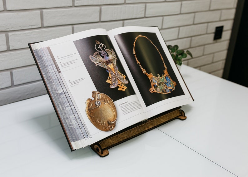 May include: An open book with a wooden stand, showcasing two pieces of jewelry. The jewelry is made of gold and features intricate designs with blue and green enamel. The book is titled "The Art Nouveau Jewelry of Ren&eacute; Lalique".