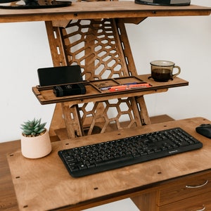 K&ouml;nnte beinhalten: Ein Stehpult aus Holz mit einer schwarzen Tastatur und Maus, einer Tasse Kaffee und einer kleinen Topfpflanze. Der Schreibtisch hat ein einzigartiges Wabenmuster und ein Regal zum Verstauen von Gegenst&auml;nden.