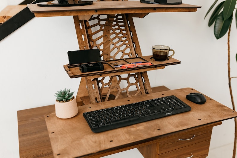 K&ouml;nnte beinhalten: Ein Stehpult aus Holz mit einer schwarzen Tastatur und Maus, einer Tasse Kaffee und einer kleinen Topfpflanze. Der Schreibtisch hat ein einzigartiges Wabenmuster und ein Regal zum Verstauen von Gegenst&auml;nden.