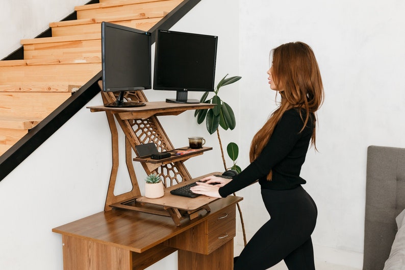 K&ouml;nnte beinhalten: Ein Stehpult aus Holz mit zwei schwarzen Computermonitoren, einer Tastatur und einer Topfpflanze. Eine Person steht am Schreibtisch und tippt auf der Tastatur.