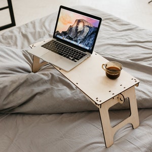 May include: A light wood laptop stand with a laptop and a cup of coffee on it. The stand is on a gray bedspread.