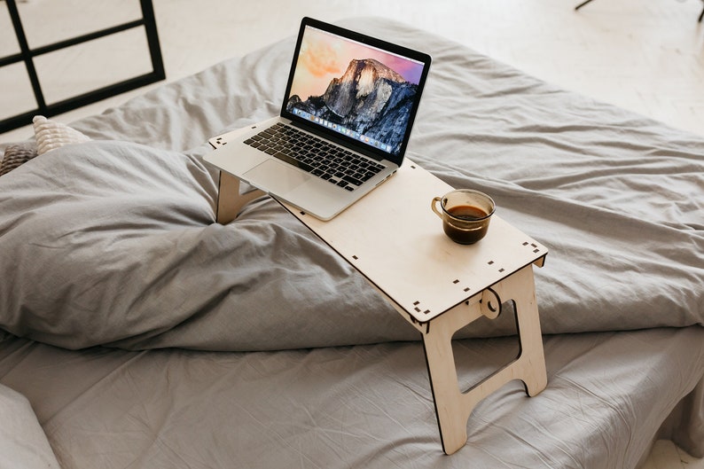 May include: A light wood laptop stand with a laptop and a cup of coffee on it. The stand is on a gray bedspread.