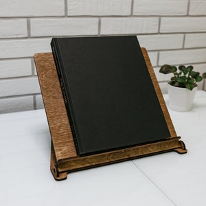 May include: A wooden book stand with a black book resting on top. The stand is made of dark wood and has a simple design. The book is closed and has a plain black cover.