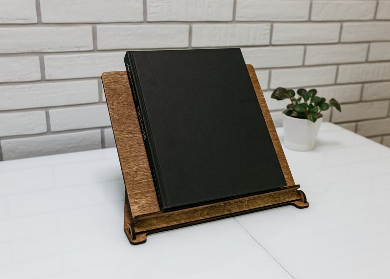 May include: A wooden book stand with a black book resting on top. The stand is made of dark wood and has a simple design. The book is closed and has a plain black cover.