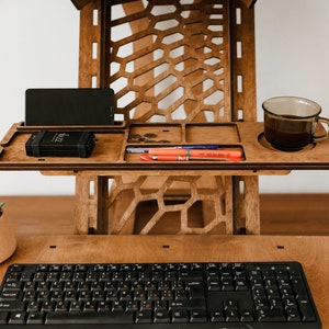 K&ouml;nnte beinhalten: Ein Stehpult aus Holz mit einem eingebauten Regal f&uuml;r ein Telefon, Stifte und eine Tasse Kaffee. Der Schreibtisch hat ein Wabenmuster und ist aus hellem braunem Holz gefertigt. Eine Tastatur und eine Maus befinden sich auf dem unteren Regal.