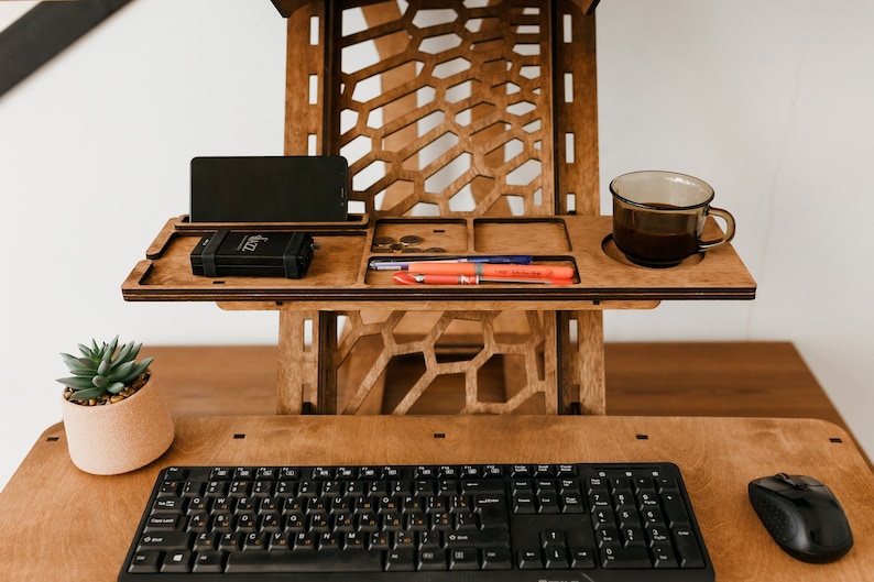 K&ouml;nnte beinhalten: Ein Stehpult aus Holz mit einem eingebauten Regal f&uuml;r ein Telefon, Stifte und eine Tasse Kaffee. Der Schreibtisch hat ein Wabenmuster und ist aus hellem braunem Holz gefertigt. Eine Tastatur und eine Maus befinden sich auf dem unteren Regal.
