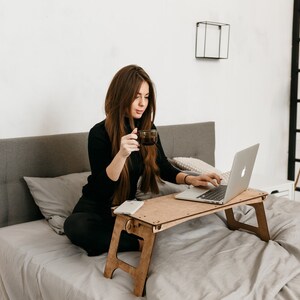 May include: A woman sits on a bed with a laptop on a wooden lap desk. She is holding a cup of coffee in one hand and typing on the laptop with the other. The bed has a gray headboard and a white blanket.