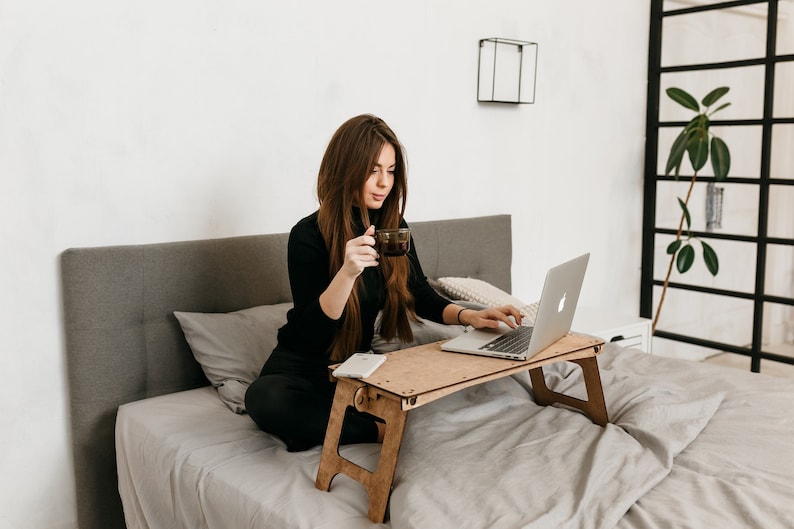 May include: A woman sits on a bed with a laptop on a wooden lap desk. She is holding a cup of coffee in one hand and typing on the laptop with the other. The bed has a gray headboard and a white blanket.