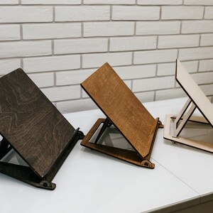 May include: Three wooden book stands in different shades of brown, natural wood, and dark brown. The stands are angled and have a simple design with a flat surface for holding books or tablets.
