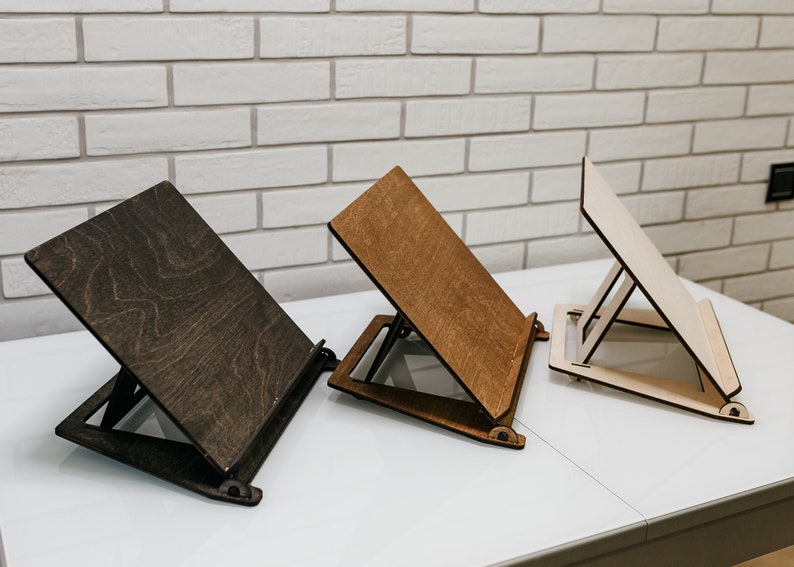 May include: Three wooden book stands in different shades of brown, natural wood, and dark brown. The stands are angled and have a simple design with a flat surface for holding books or tablets.