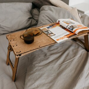 May include: A wooden folding table with a magazine and a cup of coffee on top. The table is sitting on a gray bedspread.