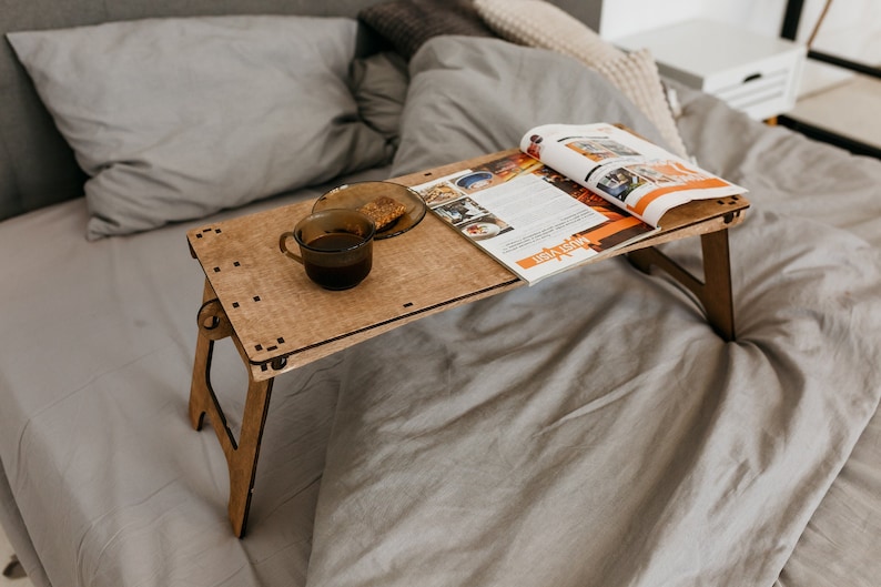 May include: A wooden folding table with a magazine and a cup of coffee on top. The table is sitting on a gray bedspread.