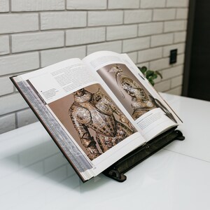 May include: An open book with illustrations of armor on a white table. The book is propped up on a black metal book stand.