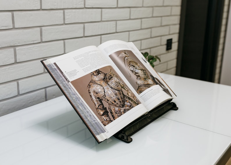 May include: An open book with illustrations of armor on a white table. The book is propped up on a black metal book stand.