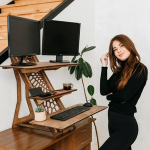K&ouml;nnte beinhalten: Ein Stehpult aus Holz mit zwei schwarzen Computermonitoren, einer Tastatur, einer Topfpflanze und einer Tasse Kaffee. Der Schreibtisch ist auf Stehh&ouml;he angehoben, und eine Frau steht daneben.