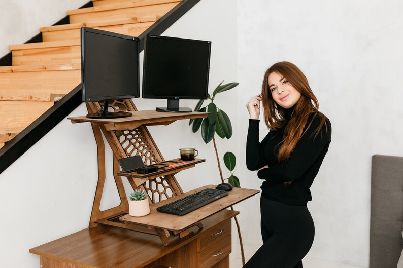 K&ouml;nnte beinhalten: Ein Stehpult aus Holz mit zwei schwarzen Computermonitoren, einer Tastatur, einer Topfpflanze und einer Tasse Kaffee. Der Schreibtisch ist auf Stehh&ouml;he angehoben, und eine Frau steht daneben.