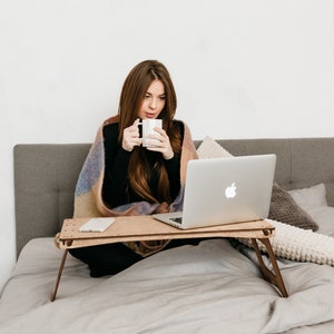 May include: A woman sits on a bed with a laptop on a wooden tray table. She is wearing a black shirt and a blanket. She is holding a white mug.