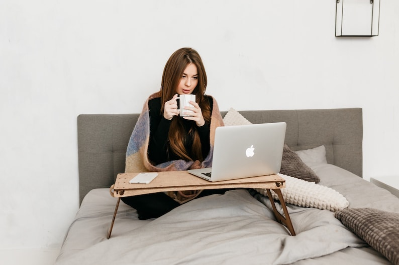 May include: A woman sits on a bed with a laptop on a wooden tray table. She is wearing a black shirt and a blanket. She is holding a white mug.