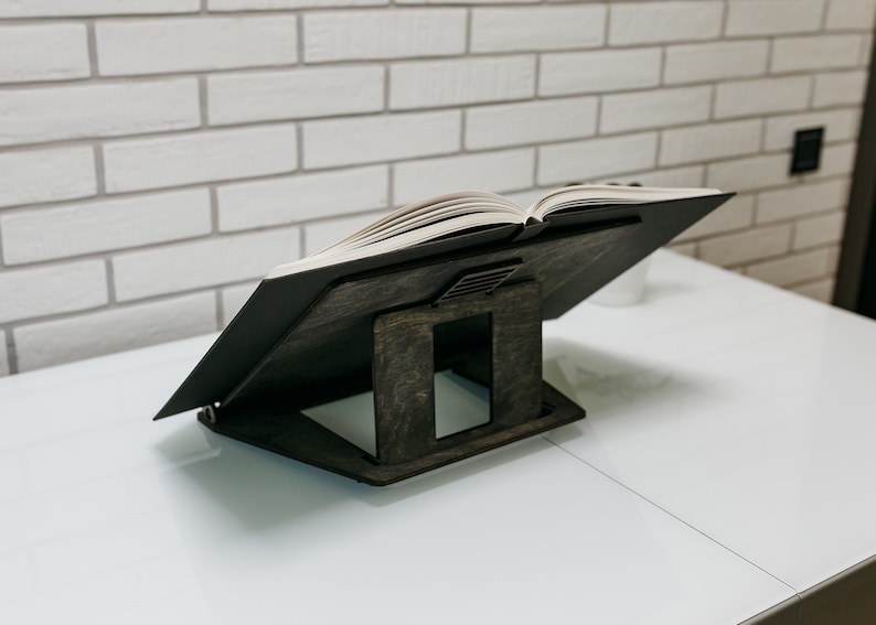 May include: A dark brown wooden book stand with a book resting on top. The stand has a unique design with a raised platform and a cutout in the back.