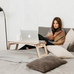 May include: A woman sits in bed with a blanket wrapped around her, using a laptop on a wooden lap desk. The bed has a gray duvet and several pillows.